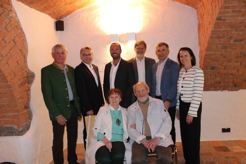 Acht Personen stehen und sitzen in einem Raum, wahrscheinlich für ein Foto. Ein Mann und eine Frau sitzen auf einem Stuhl in der Mitte. Der Raum hat weiße Wände und eine Ziegeldecke.