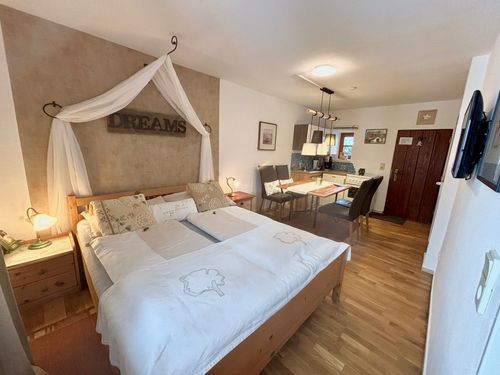 Ein Schlafzimmer mit einem Bett, Kissen und einem Baldachin mit der Aufschrift Träume. In der Nähe ein Esstisch mit Stühlen und ein Küchenbereich. Der Raum hat Holzböden und weiße Wände.
