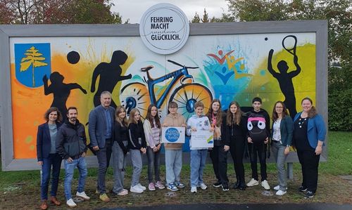 Eine Gruppe von Menschen steht vor einer Wand mit Graffiti. Sie halten Schilder und scheinen für ein Foto zu posieren. Dahinter ist eine große Graffiti mit dem Text 'FEHRING MACHT RUNDUM GLÜCKLICH'. Bäume sind im Hintergrund zu sehen.