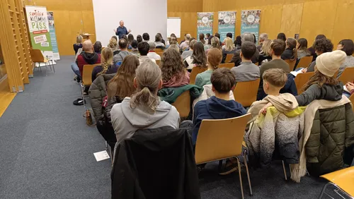 Ein Mann hält eine Präsentation in einem Klassenzimmer. Mehrere Personen sitzen vor ihm. Dahinter befindet sich eine Tafel. Das Klassenzimmer hat Holzwände und einen Teppichboden.