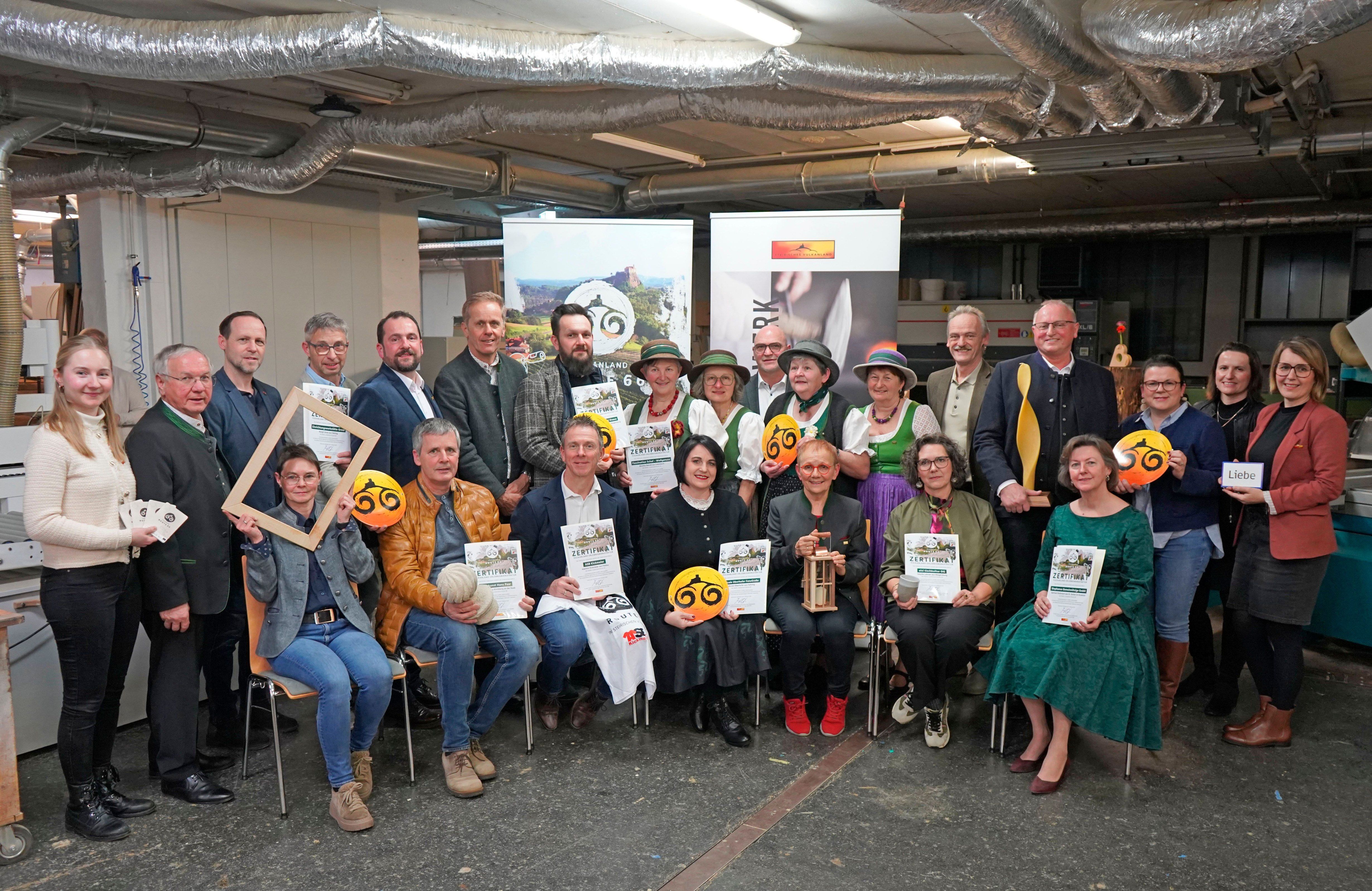 Eine Gruppe von Personen, einige sitzend und andere stehend, posiert für ein Foto mit Zertifikaten und einem Banner dahinter. Sie alle lächeln, und die Umgebung hat ein Banner, Lichter und Rohre an der Decke.