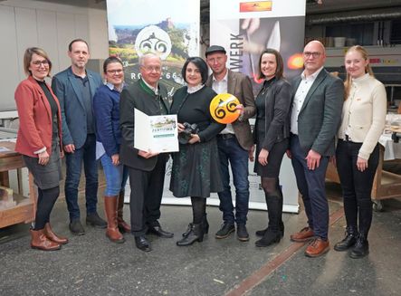 Mehrere Personen stehen in einer Reihe, wahrscheinlich bei einer Preisverleihung. Ein Mann hält ein Zertifikat und einen gelben Ball. Ein anderer Mann hält eine Kamera. Dahinter befindet sich ein Banner mit dem Wort 'WERK'.