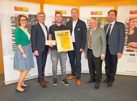 Eine Gruppe von Menschen in formeller Kleidung steht vor einem Banner. Ein Mann hält ein gerahmtes Zertifikat, während andere lächeln und zur Kamera schauen.