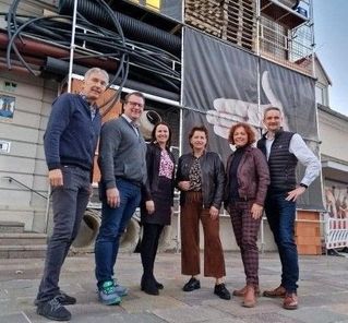 Eine Gruppe von Menschen steht vor einem Gebäude im Bau. Sie sind in Freizeitkleidung gekleidet. Ein Banner mit einem Handsymbol ist am Gebäude befestigt.