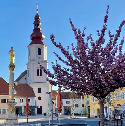Eine Kirche mit Turm und Statue steht auf einem Stadtplatz. Ein Kirschblütenbaum blüht in der Nähe, und es gibt Schirme und Bänke.
