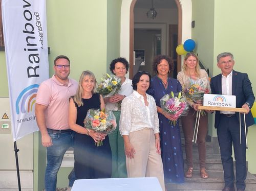 Eine Gruppe von Menschen steht auf den Stufen eines Hauses und hält Blumen. Sie lächeln und scheinen für ein Foto zu posieren. Hinter ihnen ist ein Banner mit dem Text 'LebensGroB' und Luftballons.