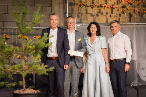 Vier Personen stehen lächelnd nebeneinander, einer hält ein Zertifikat. Sie stehen vor einer Wand mit Sonnenblumen in Vasen.