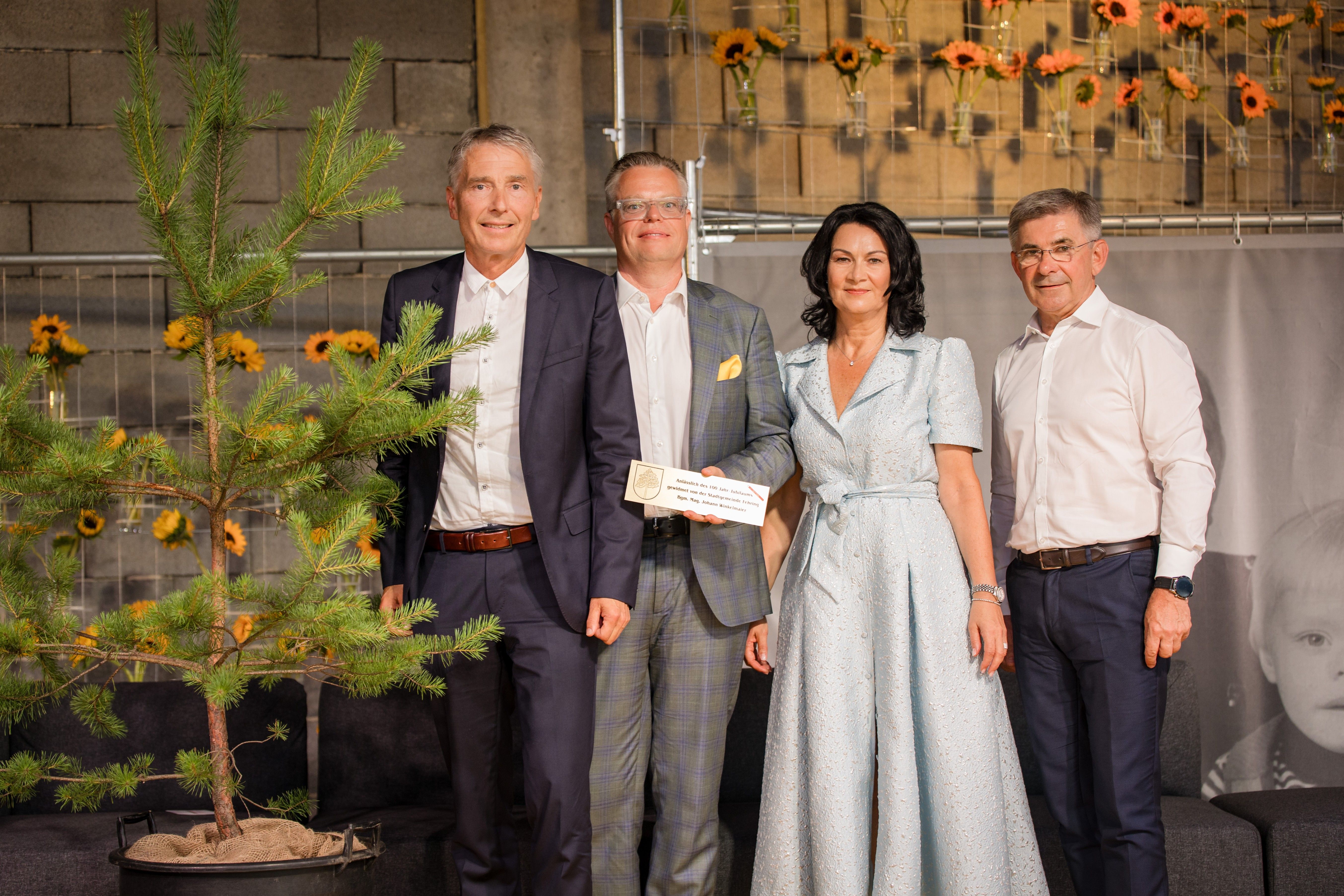 Vier Personen stehen lächelnd nebeneinander, einer hält ein Zertifikat. Sie stehen vor einer Wand mit Sonnenblumen in Vasen.