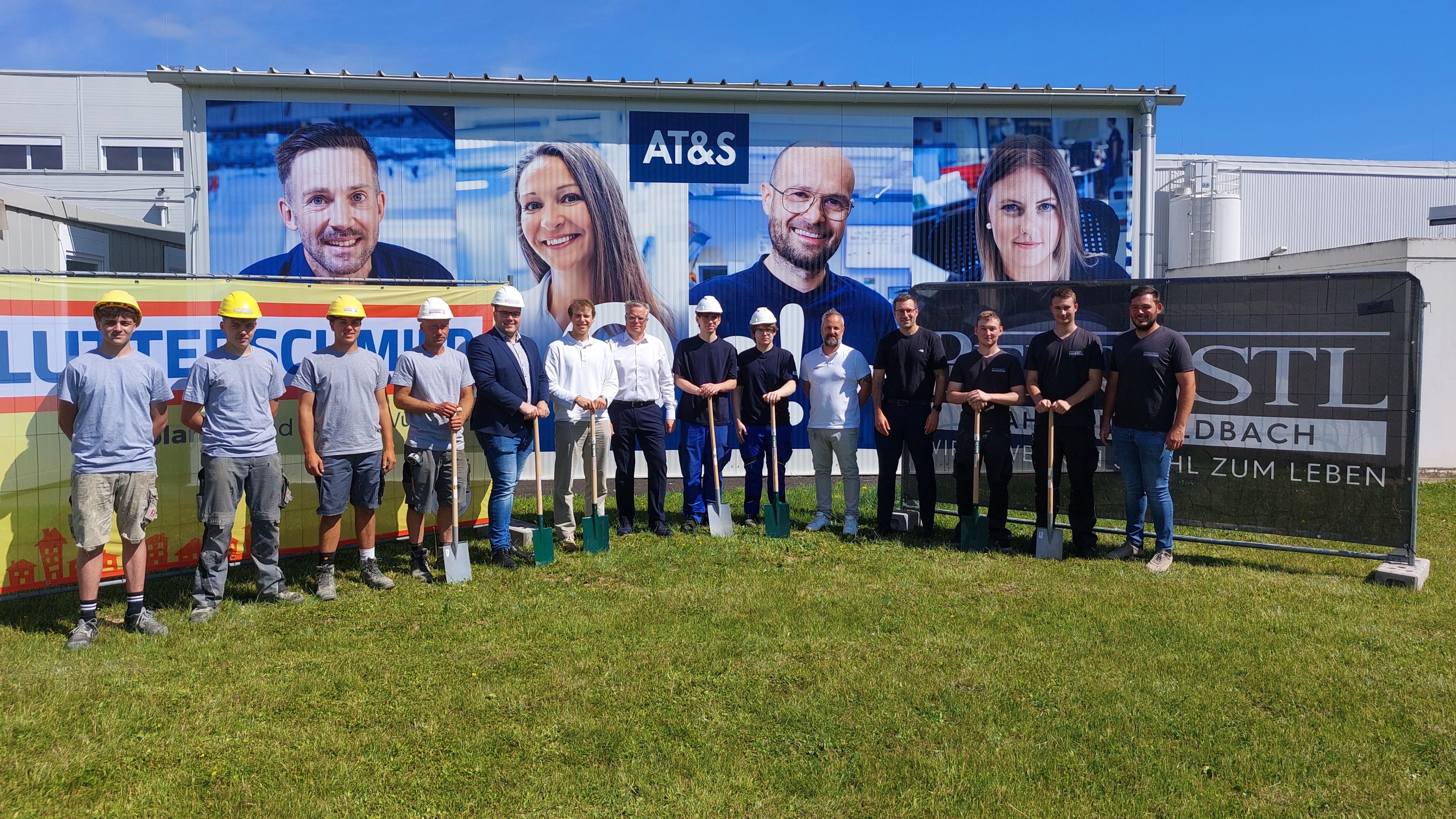 Eine Gruppe von Menschen in Bauausrüstung posiert für ein Foto vor einem großen Banner mit AT&S-Logo und lächelnden Gesichtern.