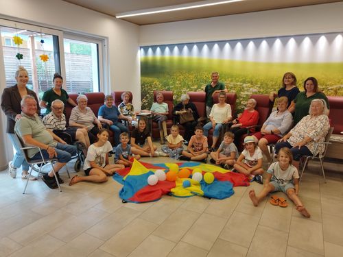 Eine Gruppe älterer Menschen und Kinder sitzt in Stühlen und auf dem Boden um ein farbenfrohes Fallschirm mit Luftballons.