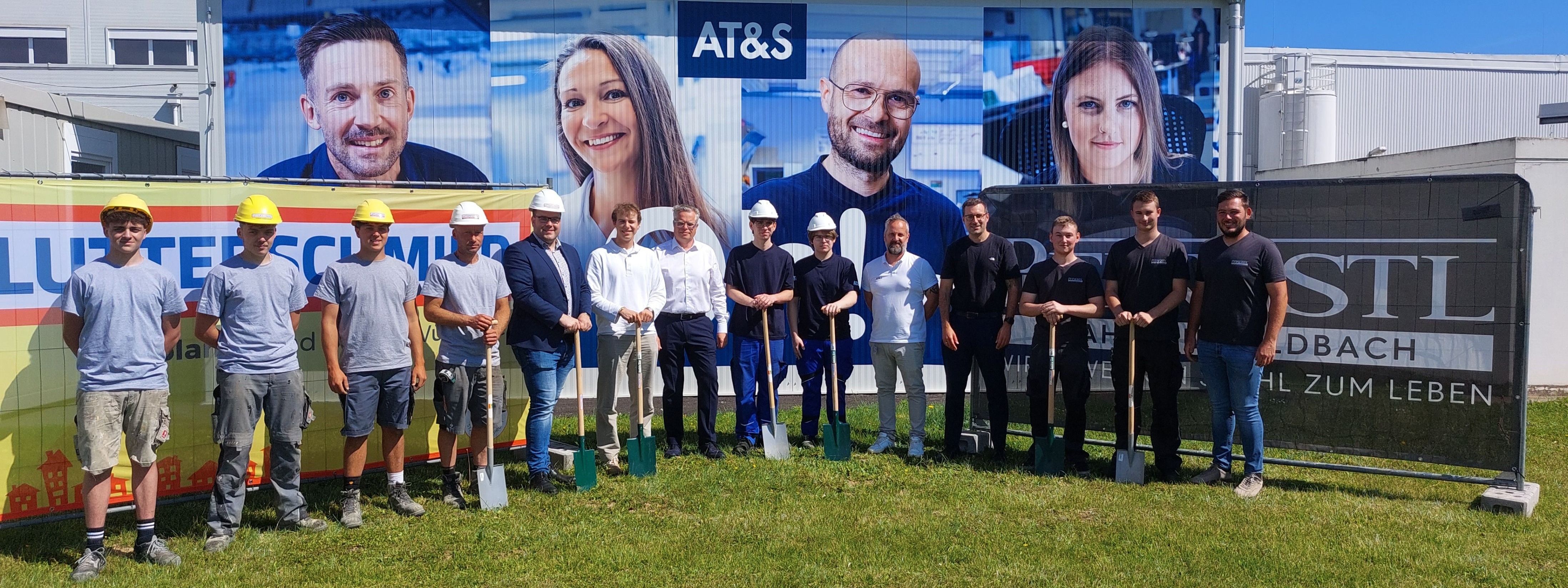 Eine Gruppe von Menschen in Helmen und mit Schaufeln posiert für ein Foto vor einer großen AT&S-Werbung.