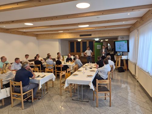 Eine Gruppe von Menschen sitzt an Tischen in einem Konferenzraum, mit zwei Männern, die an einem Podium vor einem Bildschirm stehen. Der Raum hat Holzbalken und weiße Wände.