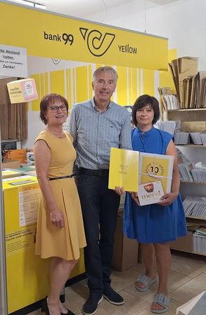 Drei Personen stehen vor einem gelben Hintergrund mit dem Text 'bank 99 yellow'. Die Frau links trägt ein gelbes Kleid, der Mann in der Mitte ein blaues Hemd und die Frau rechts hält eine gelbe Mappe mit dem Text 'merci'. Sie alle lächeln. Dahinter befindet sich ein Regal mit mehreren Büchern.