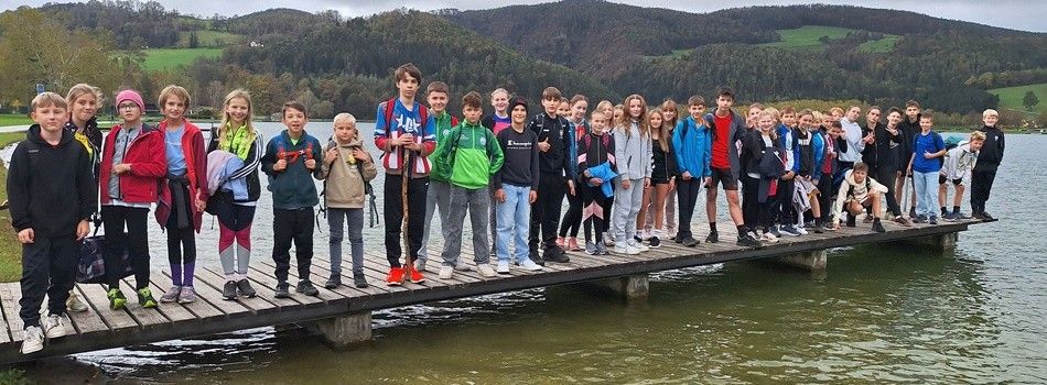 Eine Gruppe junger Menschen steht auf einem Holzsteg am See, einige tragen Rucksäcke und halten Stöcke.