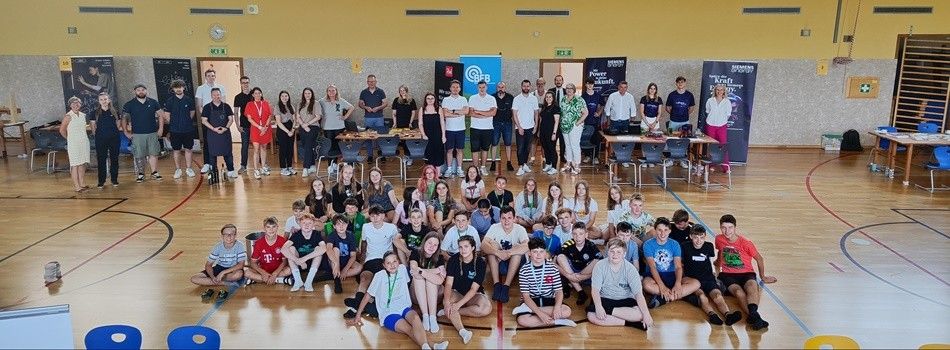 Eine Gruppe von Menschen, sowohl Erwachsene als auch Kinder, versammelt sich in einer Turnhalle für ein Foto. Einige sitzen auf dem Boden, während andere hinter Tischen stehen. An den Wänden sind Banner zu sehen.