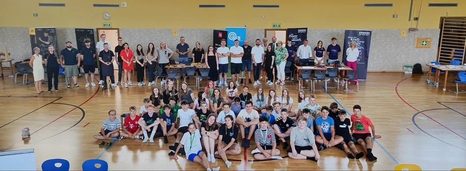 Eine Gruppe von Menschen, sowohl Erwachsene als auch Kinder, versammelt sich in einer Turnhalle für ein Foto. Einige sitzen auf dem Boden, während andere hinter Tischen stehen. An den Wänden sind Banner zu sehen.