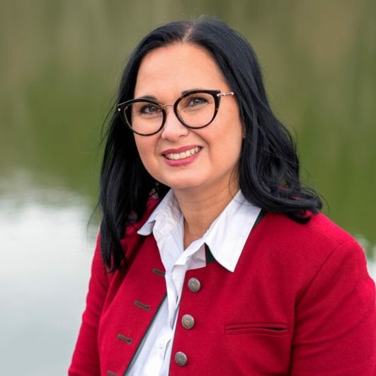Eine Frau mit schwarzen Haaren, die eine Brille und eine rote Jacke trägt, lächelt und steht in der Nähe eines Gewässers.