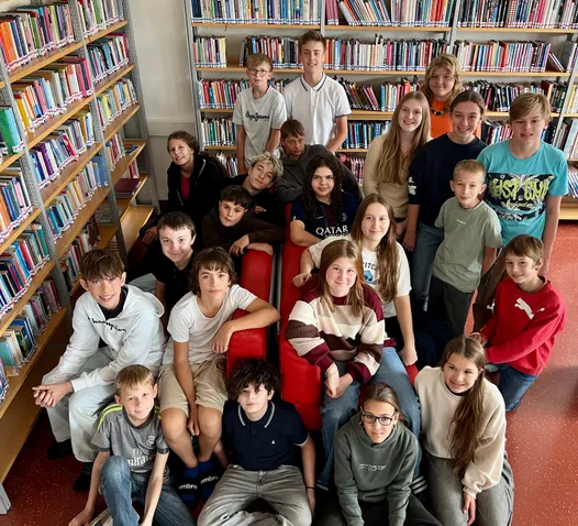 Eine Gruppe junger Schüler posiert für ein Foto in einer Bibliothek. Sie sitzen auf roten Stühlen und hinter ihnen stehen Regale voller Bücher.