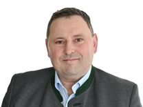 Ein Mann in einem grauen Anzug mit grünem Kragen lächelt für ein Porträt. Er trägt ein blaues Hemd und hat kurze Haare.