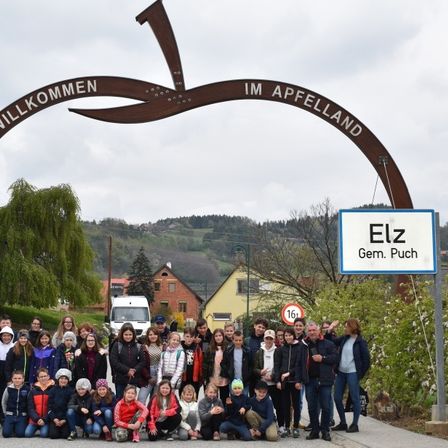 Eine Gruppe von Menschen steht unter einem Schild, das Willkommen IM Apfelland steht, in der Nähe eines Ortsschilds für Elz Gem. Puch. Sie alle lächeln und posieren für ein Foto.