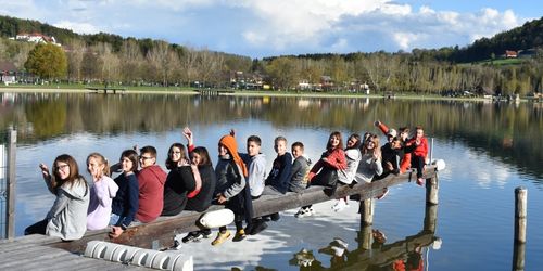 Eine Gruppe junger Menschen sitzt auf einem Steg am See, lächelt und posiert für ein Foto. Der See ist von Bäumen und Gebäuden umgeben.