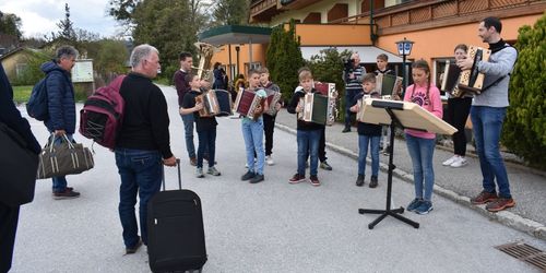 Eine Gruppe von Kindern und Erwachsenen spielt Musikinstrumente im Freien. Die Kinder spielen Akkordeons, während ein Erwachsener eine Tuba spielt. Sie stehen auf der Straße vor einem Gebäude.