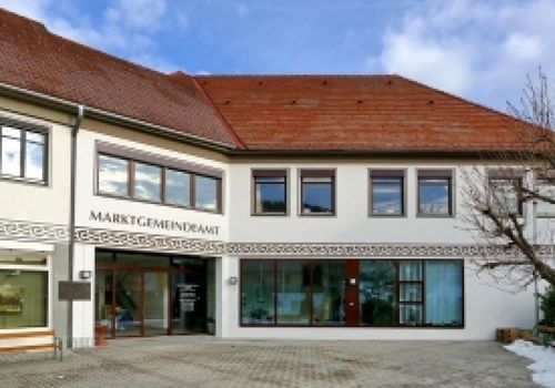 Ein weißes Gebäude mit einem roten Dach und großen Fenstern. Der Name 'MARKTGEMEINDEAM' steht auf der Vorderseite.