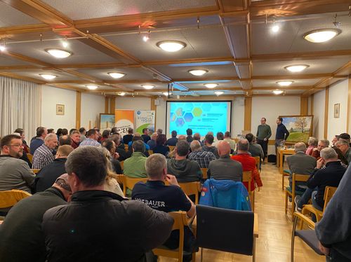 Ein Konferenzraum mit Menschen, die einer Präsentation folgen. Eine große Leinwand zeigt ein Sechseck-Design, und zwei Männer stehen vorne. Das Publikum sitzt auf Stühlen, einige tragen Jacken. Der Raum hat Holzwände und einen Holzboden.