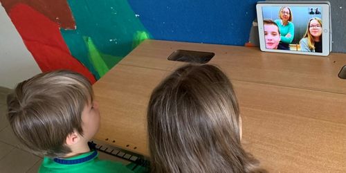 Ein Junge und ein Mädchen sitzen an einem Schreibtisch und beobachten einen Videoanruf auf einem Tablet. Eine Frau erscheint auf dem Bildschirm.