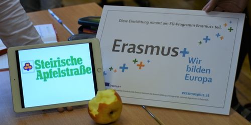Ein iPad zeigt eine Website an, während ein Erasmus+-Poster neben einem Apfel auf einem Tisch steht.