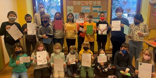 Ein Klassenzimmer voller Kinder, die Masken tragen und Papiere hochhalten, mit zwei Erwachsenen im Hintergrund. Die Wand hat Lehrposter und einen Bulletin-Board mit bunten Dekorationen.