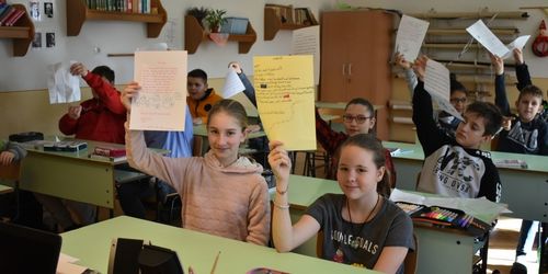 Klassenzimmerszene mit Schülern, die bunte Papiere hochhalten, Bücherregalen und einem Lehrerschreibtisch im Hintergrund.