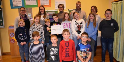 Eine Gruppe von Kindern und Erwachsenen steht in einem Klassenzimmer, lächelt und posiert für ein Foto. Ein Zertifikat mit Erasmus+ ist sichtbar.