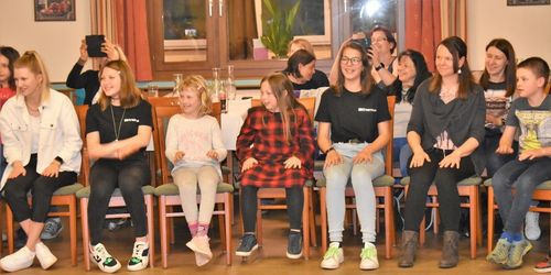 Eine Gruppe von Kindern und Erwachsenen sitzt in Stühlen, möglicherweise für ein Foto. Sie tragen Freizeitkleidung. Dahinter befinden sich Tische mit Gläsern und einer Pflanze. Zwei Personen halten Kameras.