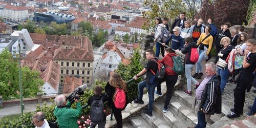 Eine Gruppe von Menschen, einige mit Rucksäcken, sind auf Stufen in der Nähe einer Stadtansicht, wobei eine Person filmt.