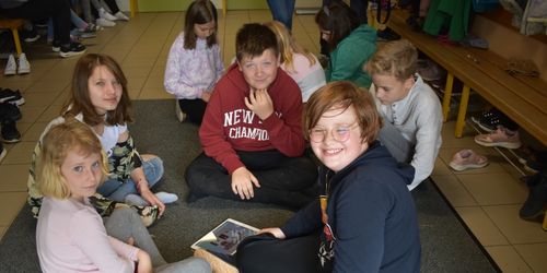 Mehrere Kinder sitzen im Kreis auf dem Boden, zwei davon mit Brille. Ein Junge trägt ein rotes Sweatshirt mit weißen Buchstaben. Ein Mädchen hält ein Tablet.