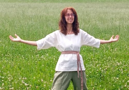 Eine Frau in einem weißen Oberteil und grüner Hose steht mit ausgestreckten Armen in einem grünen Feld und lächelt.