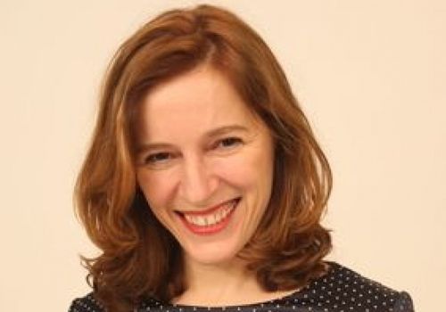 Eine Frau mit braunen gewellten Haaren und einem lächelnden Gesicht trägt ein schwarz gepunktetes Shirt. Sie schaut nach vorne und scheint für ein Foto zu posieren.