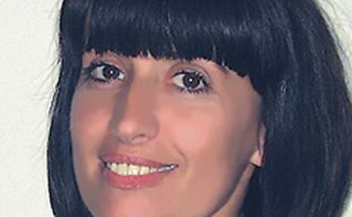 Eine Frau mit kurzen Fransen lächelt und zeigt ihre Zähne und einen leichten Muttermal auf der Wange. Sie hat braune Augen und dunkles Haar.