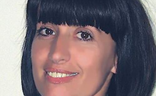 Eine Frau mit kurzen Fransen lächelt und zeigt ihre Zähne und einen leichten Muttermal auf der Wange. Sie hat braune Augen und dunkles Haar.