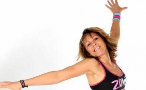 Eine Frau in Trainingskleidung lächelt und streckt ihre Arme nach oben, möglicherweise in einem Zumba-Kurs.