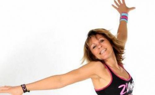 Eine Frau in Trainingskleidung lächelt und streckt ihre Arme nach oben, möglicherweise in einem Zumba-Kurs.
