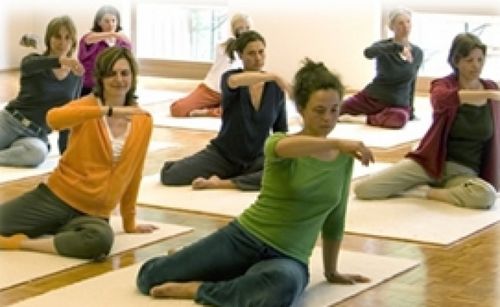 Eine Yogastunde mit Frauen auf Matten, die Posen üben und sich dehnen. Helles Zimmer mit großen Fenstern.