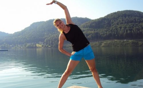 Eine Frau in einem schwarzen Tanktop und blauen Shorts dehnt sich auf einer Pier, mit einem ruhigen See und Bergen im Hintergrund.