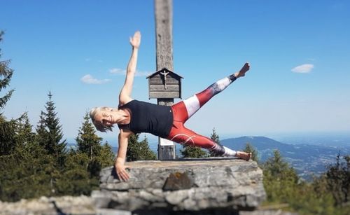 Eine Frau in Sportbekleidung praktiziert Yoga auf einem felsigen Hügel, nahe eines hölzernen Kreuzes. Dahinter stehen Bäume und man hat einen klaren Blick auf die Berge.