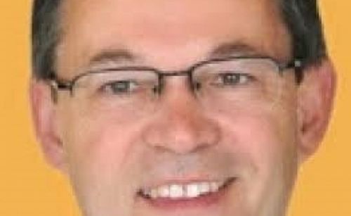 Ein Mann mit Brille lächelt mit sichtbaren Zähnen vor einem gelben Hintergrund.