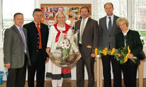 Eine Gruppe von formell gekleideten Menschen steht in einem Raum, lächelt und hält Blumen. Eine Frau in der Mitte hält einen Korb mit Blumen und einem Schild. Hinter ihnen befindet sich ein Aushang mit verschiedenen Bildern.