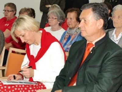 Eine Frau mit einem roten Schal schreibt auf Papier, während sie in einem Raum mit anderen Leuten sitzt. Ein Mann in Anzug und Krawatte sitzt neben ihr.