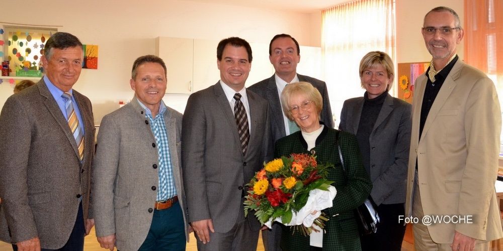 Eine Gruppe formell gekleideter Personen steht in einem Raum. Eine Frau hält einen Blumenstrauß.
