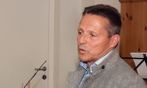 Ein Mann steht mit ernster Miene in der Nähe einer weißen Tür. Er trägt eine graue Jacke und ein blaues Hemd. Der Hintergrund ist unscharf.